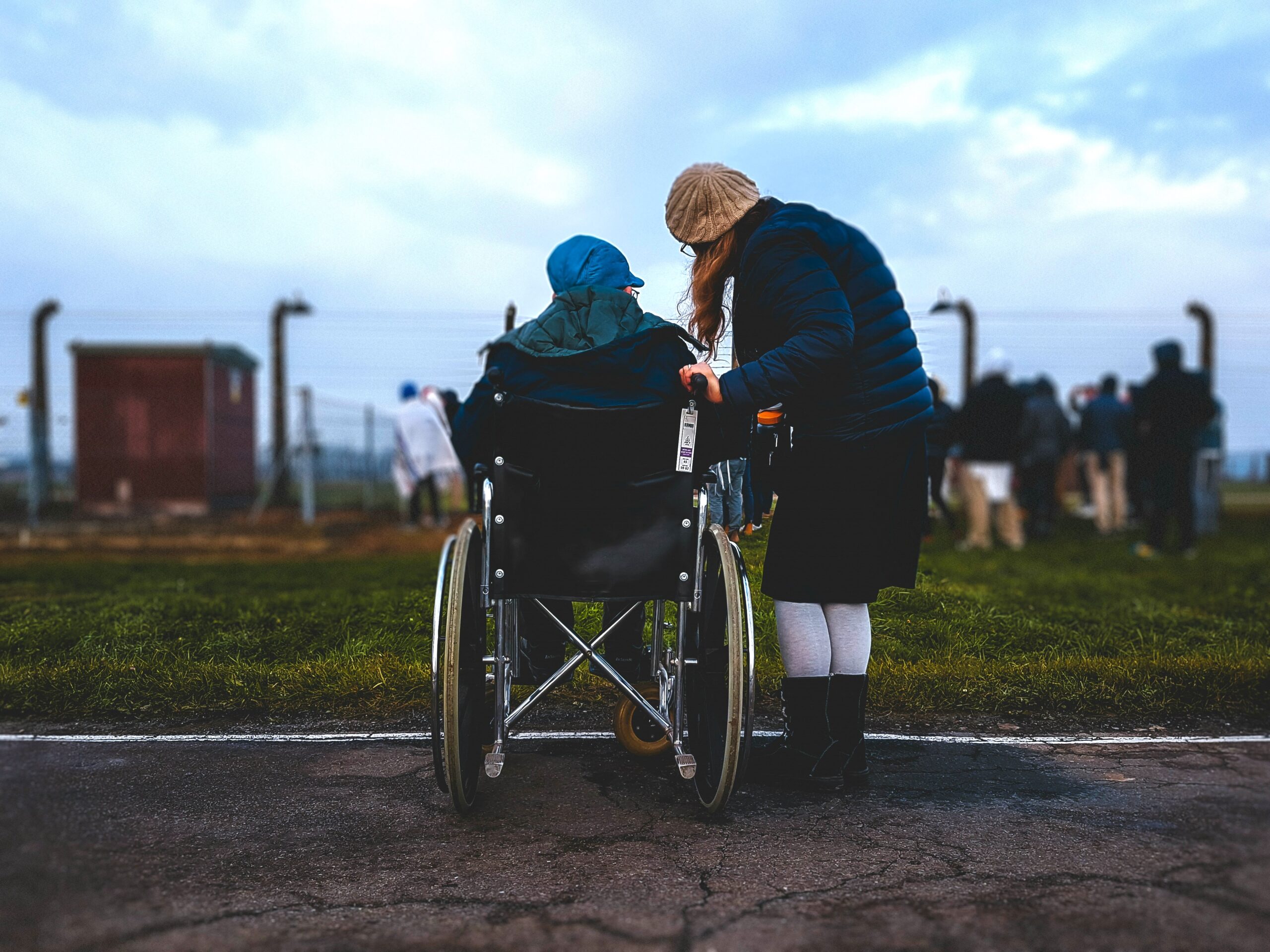 Two people, one in a wheelchair, at the edge of tarmac path. They are looking towards a crowd further away who are looking through a fence.