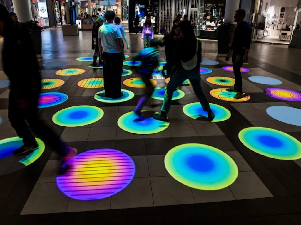 Photo of people jumping on rainbow coloured lights on the floor in a dark shopping mall.
