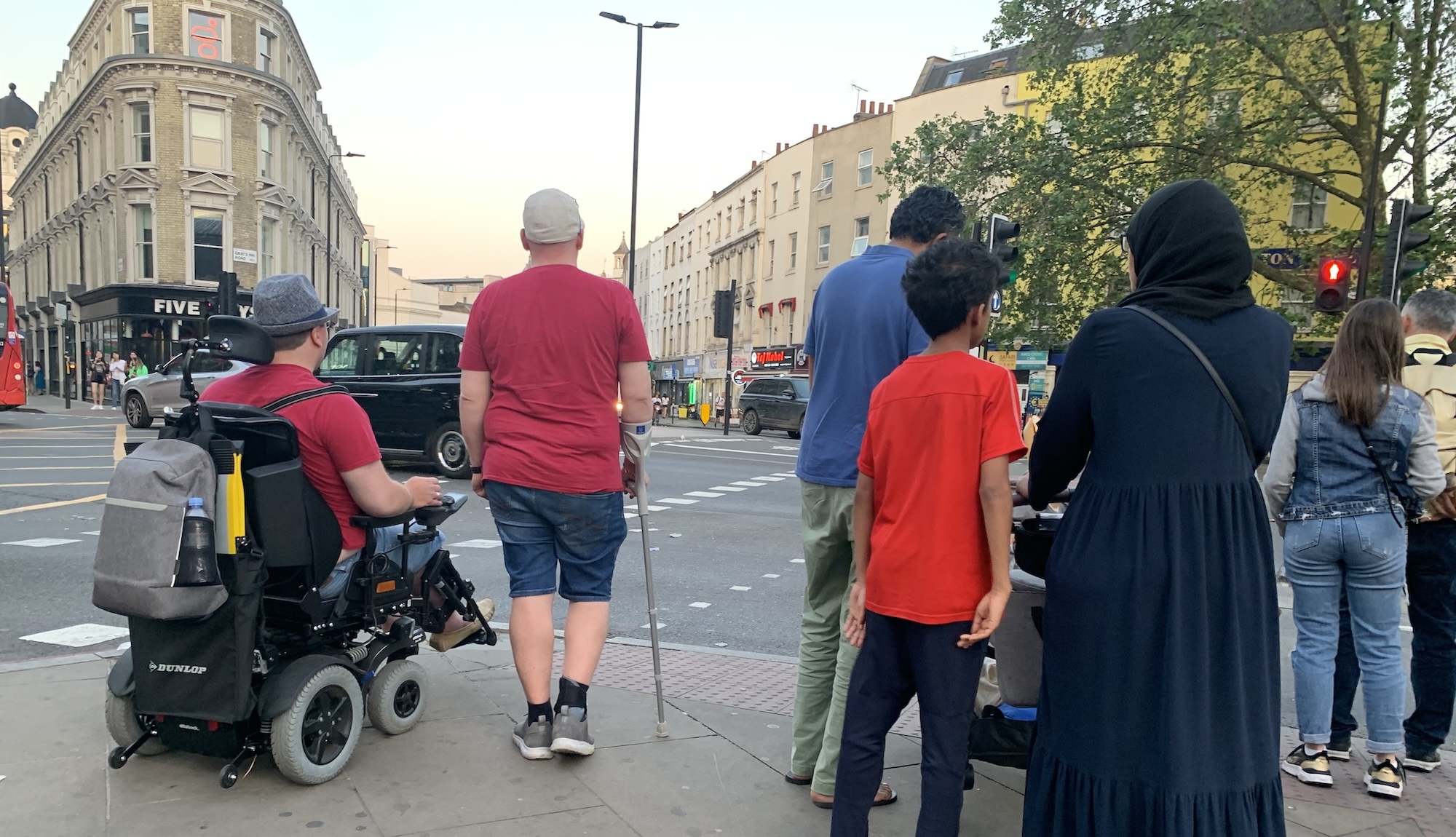A variety of people waiting to cross a busy road, some with access supports - a pram, walking stick, wheelchair.